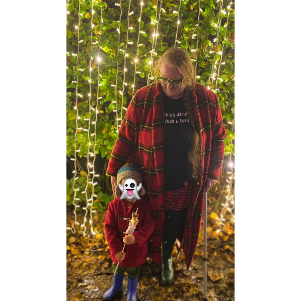 plus size woman in tartan coat with toddler in red duffle stunting infront of twinkly light wall