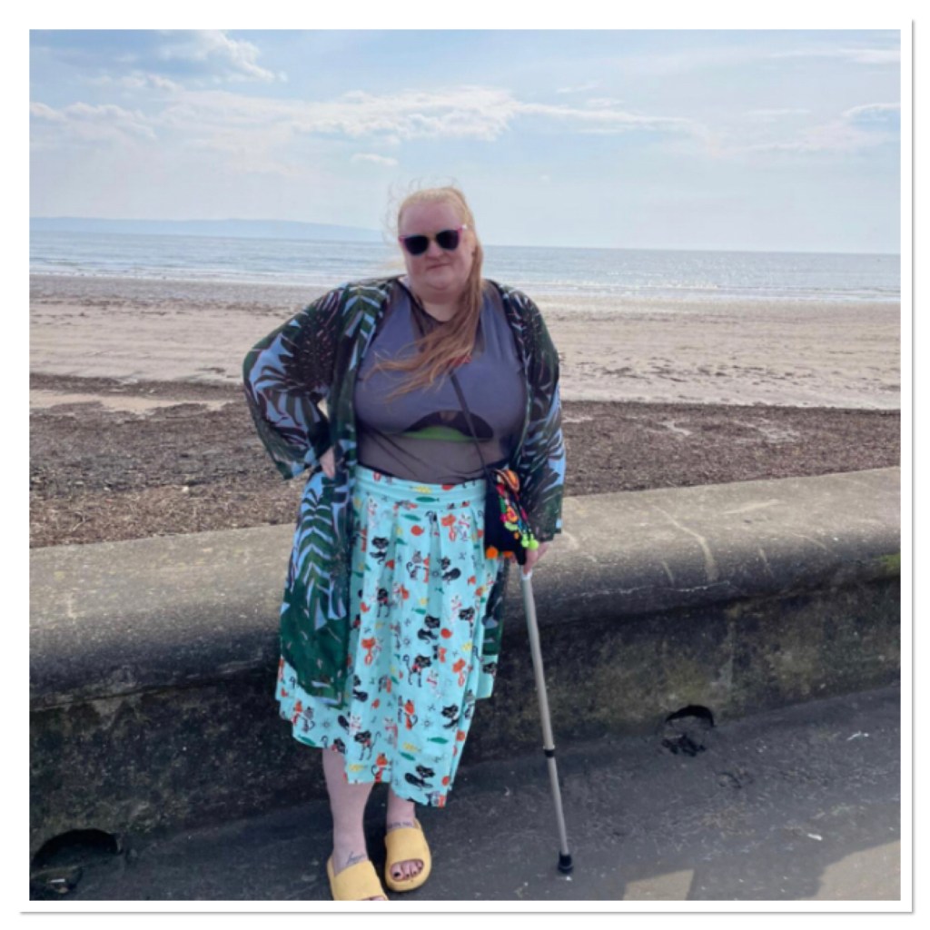 ly is standing by wall with beach in background. She is leaning on acealking stick wearing cat print swing skirt, sheer t shirt &  sunglasses