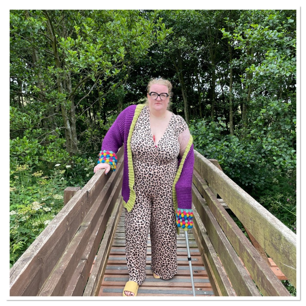 ly is wearing a leopard print jumpsuit & purple cardi.  She is standing on a wooden bridge with a walking stick 