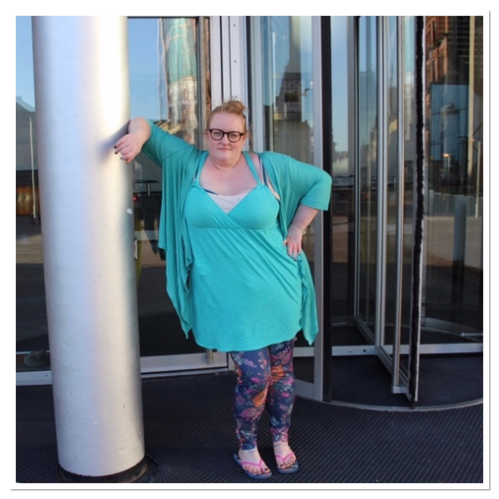 Ly  is wearing an aqua dress with floral leggings leaing against a post with a hand on her hip n