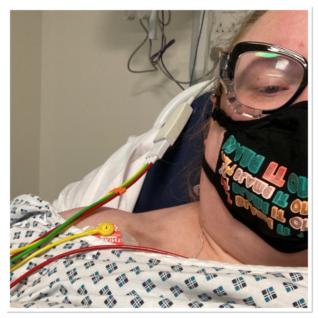 ly is in a hospital bed wearing a face mask and hospital gown. She has leads attached to her chest 