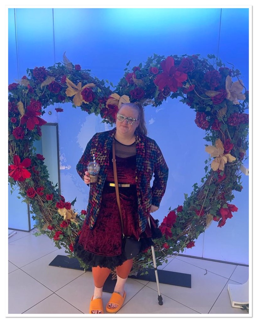 ly is standing in front of a floral heart wearing a burgundy velvet dress and blazer. She is holding a frozen coke and walking stick. 