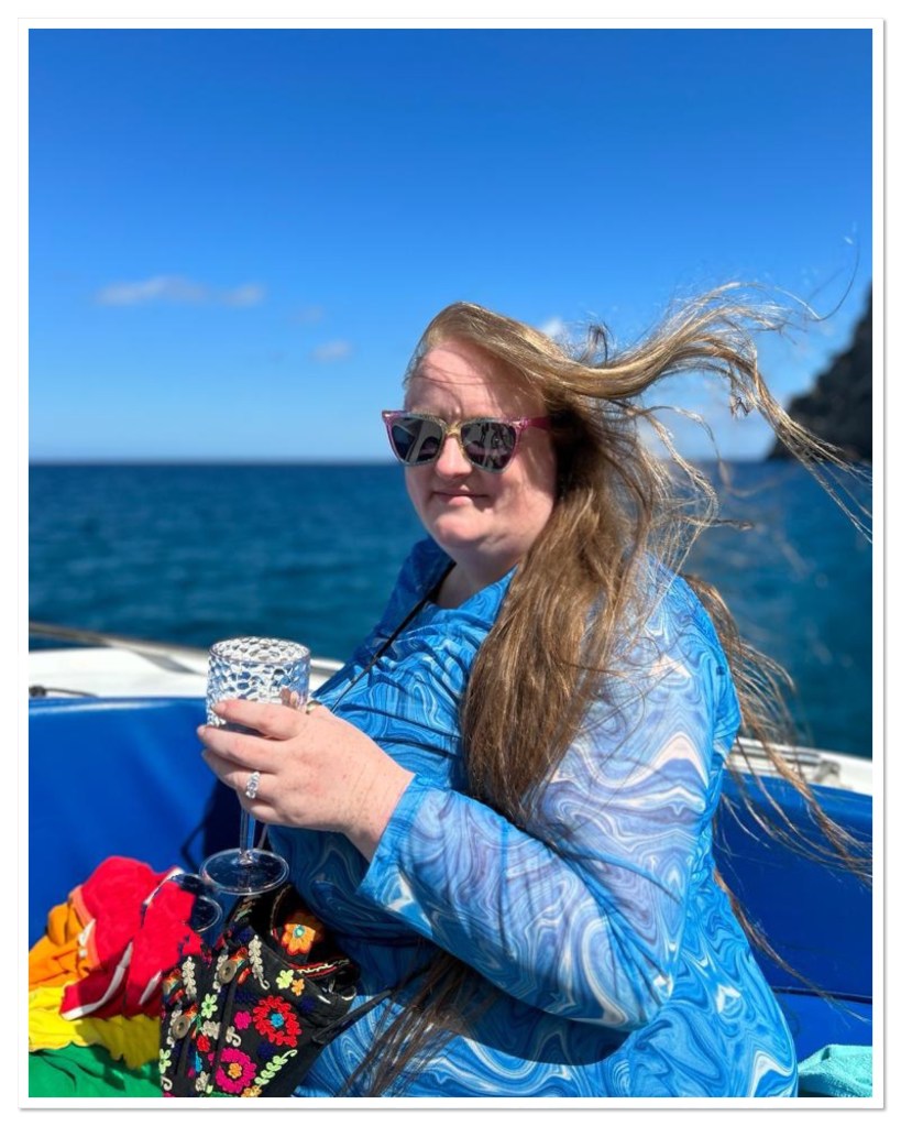 ly is wearing a blue dress and sunglasses. She is sat in boat with cliffs and ocean behind her. Her hair is blowing in the wind and she is holding a glass of rose. 
