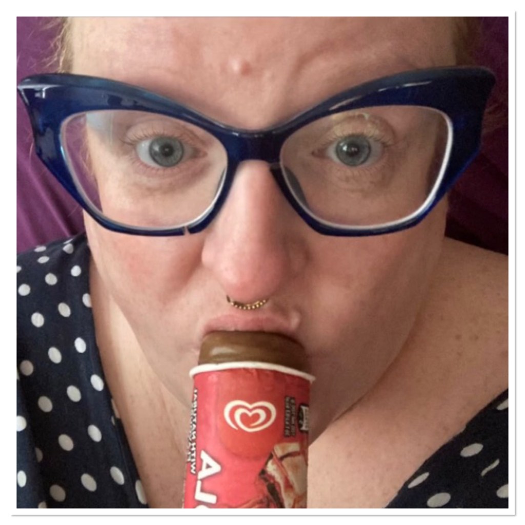 Close of ly eating an ice lolly. She is wearing blue cat eye glasses and a gold septum ring. 